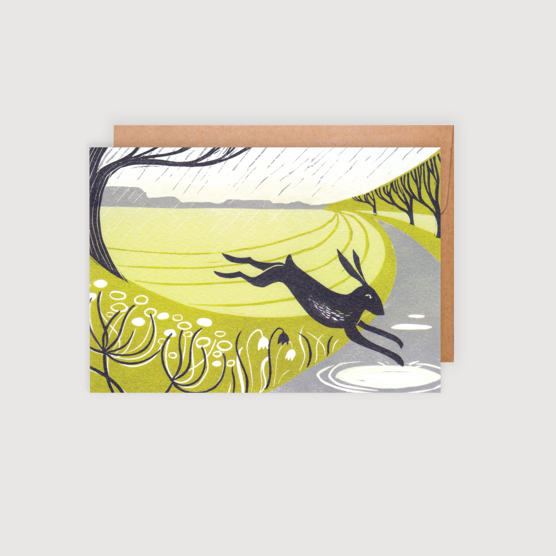 A greeting card featuring a lino print illustration of a stylized hare leaping over hedgerows, with a mix of warm sunshine and rain depicted in the background.