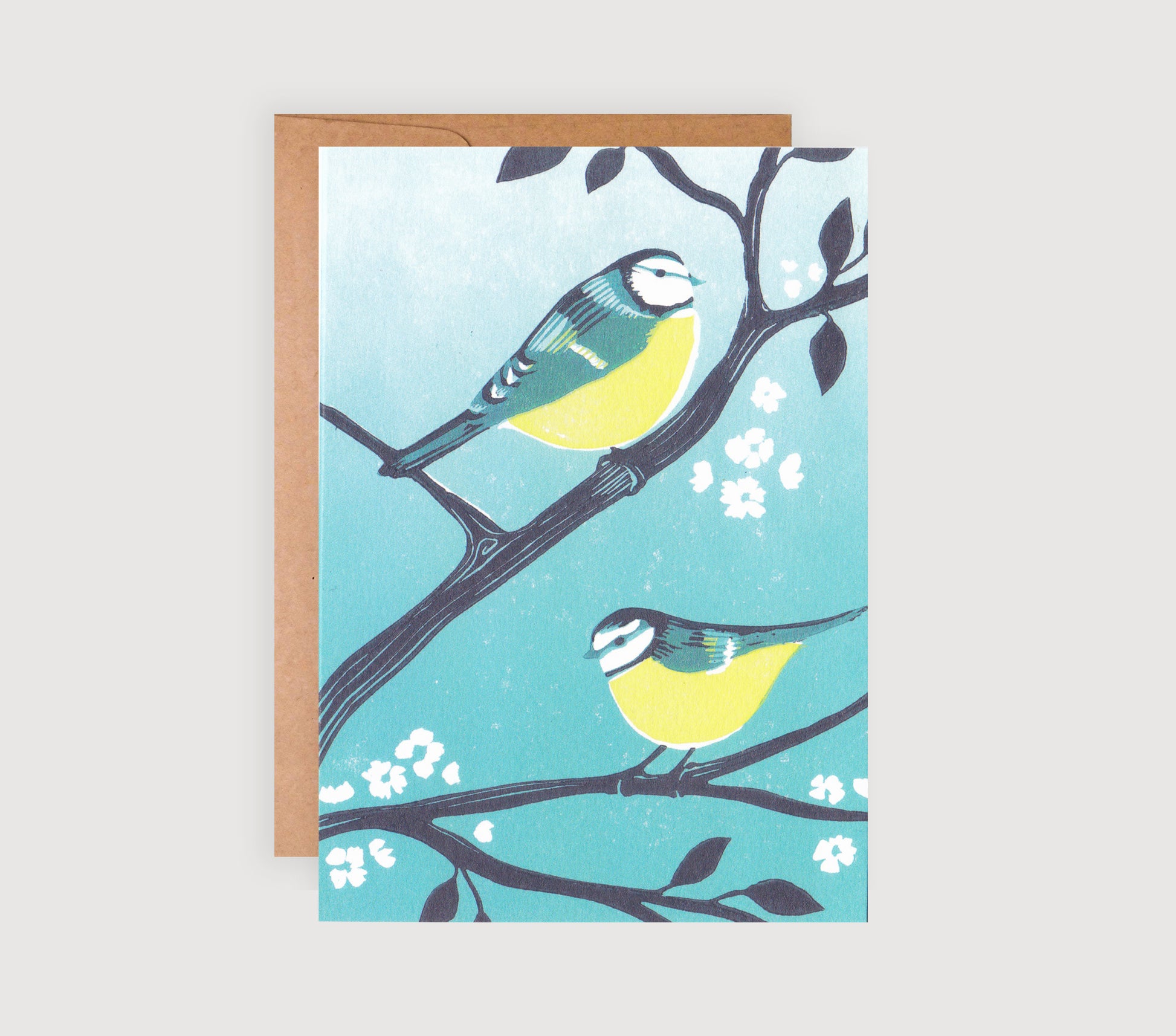 A greetings card with a linocut print of two blue tits perched on branches with turquoise background and white flowers.