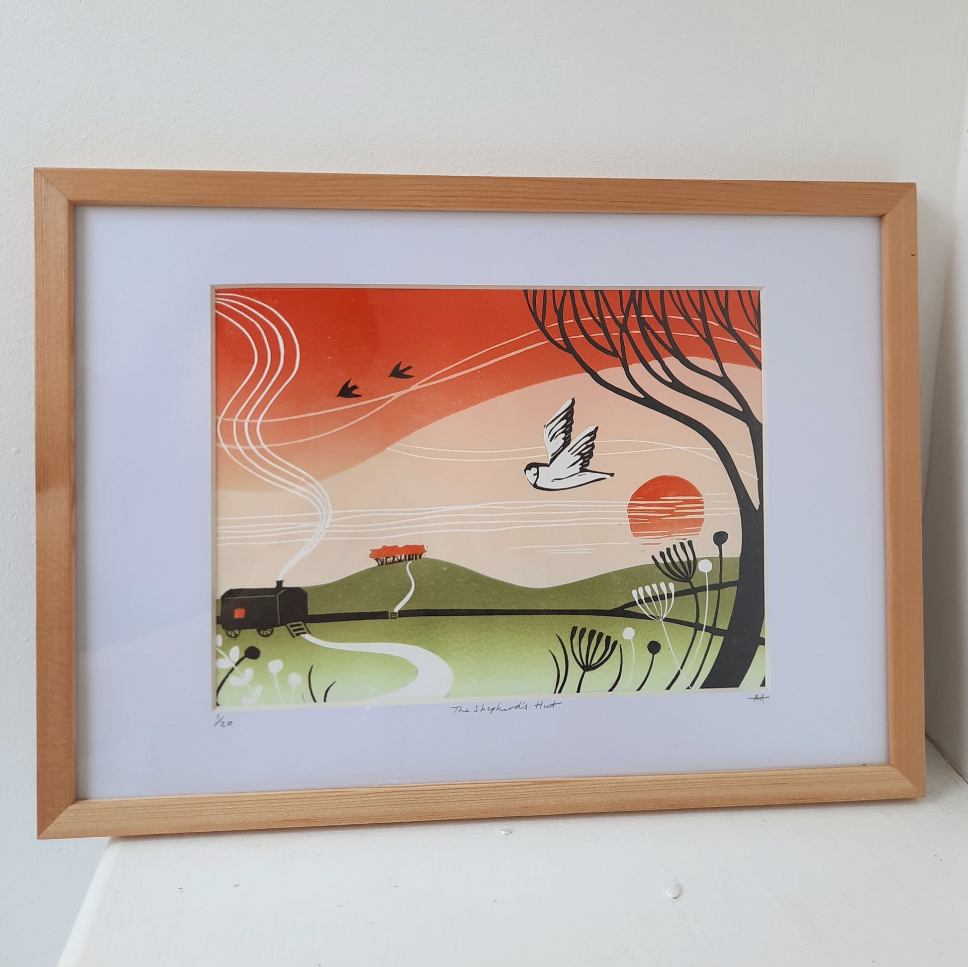 An original framed lino print featuring a countryside scene with a shepherds hut, birds, and a sunset, in a wooden frame.