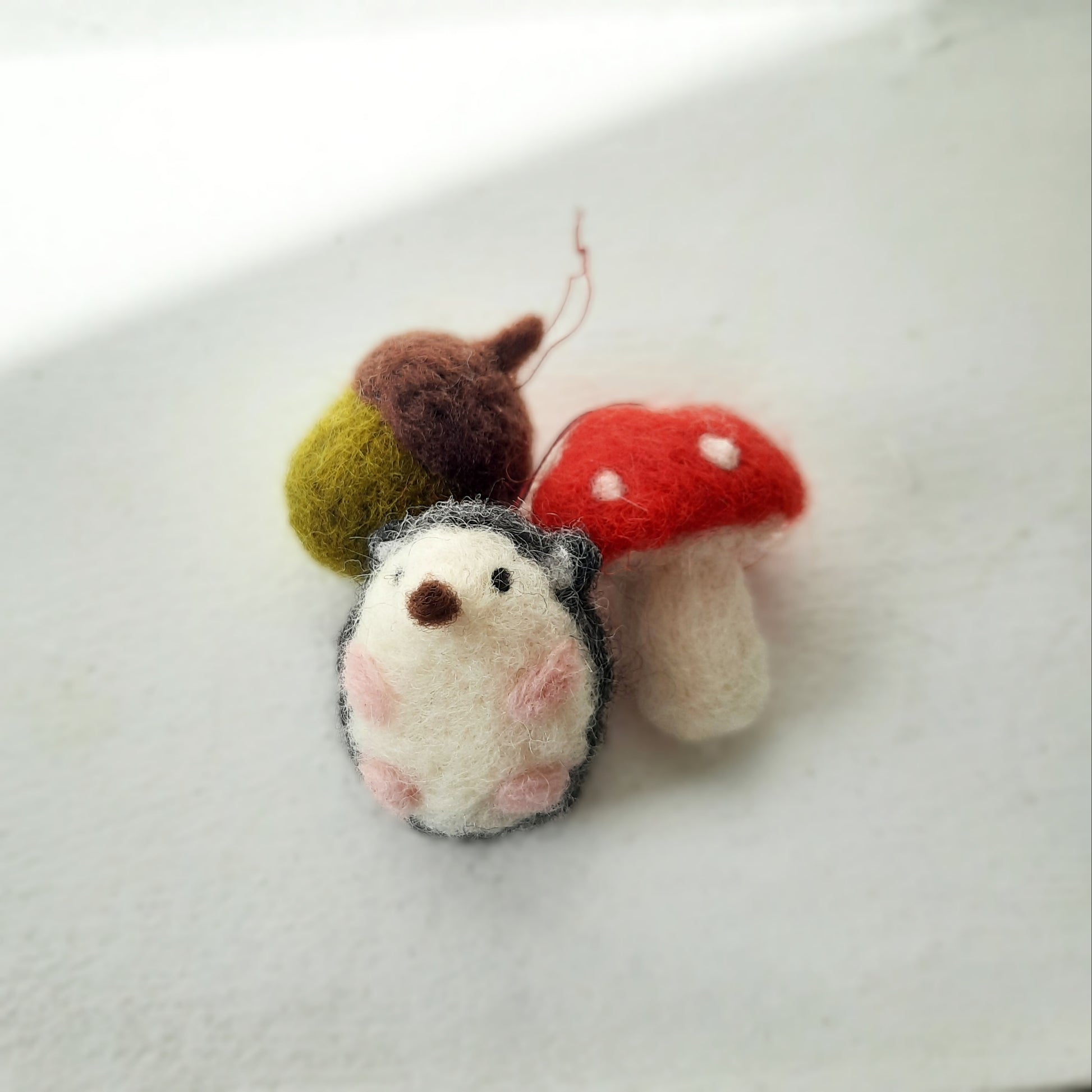 A set of three handmade needle felted decorations, including an acorn, a hedgehog, and a mushroom, in Autumnal colors.