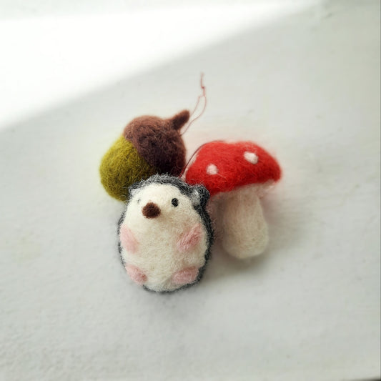 A set of three handmade needle felted decorations, including an acorn, a hedgehog, and a mushroom, in Autumnal colors.