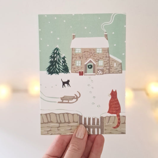 Hand holding a Christmas-themed card with a house, tree, and cats on a light background.
