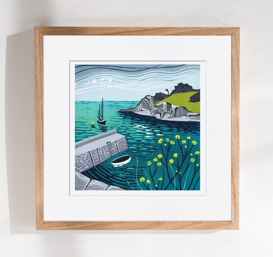 Framed artwork of a coastal scene with a sailboat, cliffs, and a building.