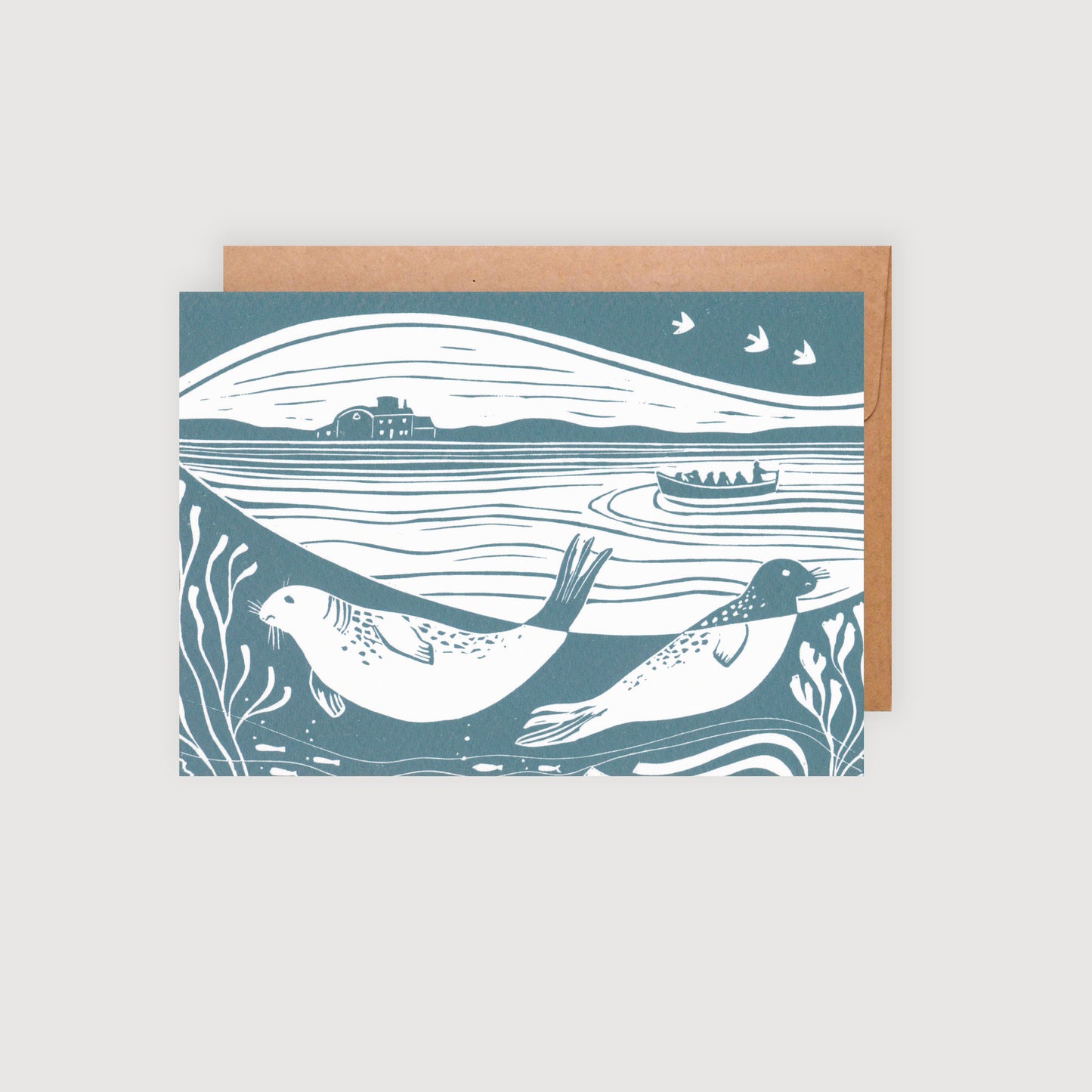An A6 greetings card featuring a lino print design with seals, birds, and a coastal landscape, in shades of blue and white, accompanied by a brown kraft envelope.