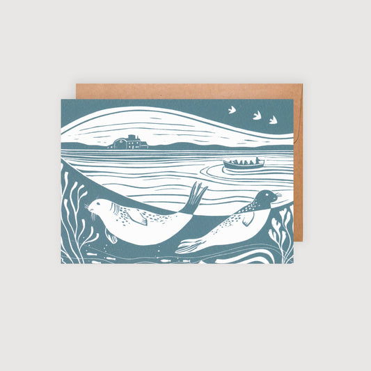 An A6 greetings card featuring a lino print design with seals, birds, and a coastal landscape, in shades of blue and white, accompanied by a brown kraft envelope.