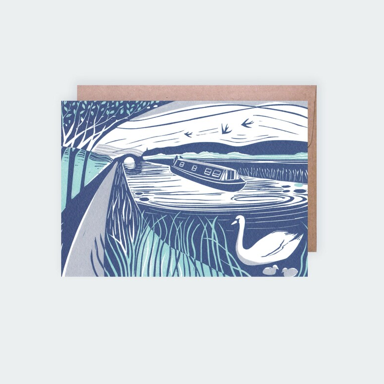 A greetings card featuring a lino print with a canal scene, including a boat, trees, birds, and a swan, on textured card stock, accompanied by a brown kraft envelope.
