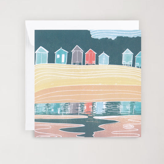 A greetings card with a linocut print featuring colorful beach huts at Wells, reflecting in water, with a blue sky above.