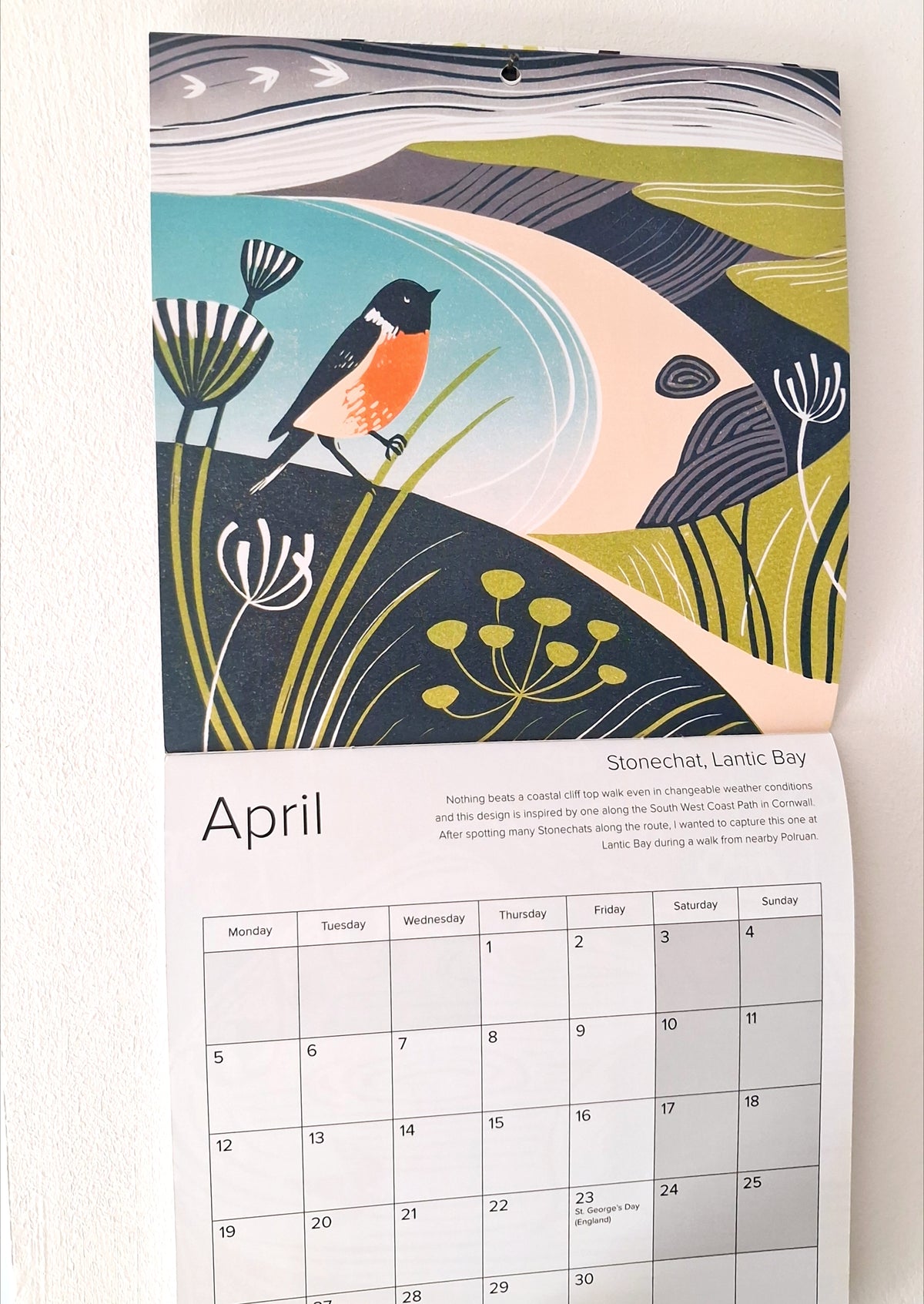 A hanging wall calendar page showing April with a stylized nature illustration above it. The artwork features a bird over water and grasses in muted teal, cream, olive, navy, and orange tones.